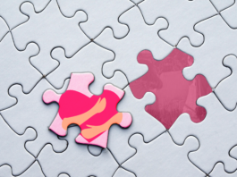 A puzzle with one missing piece. The remaining piece is pink with a red heart and two hands.