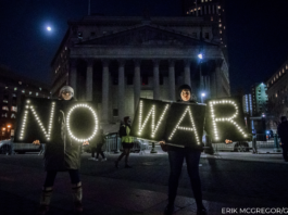 An illuminated "No War" sign shines in the darkness. Two protesters are holding the sign.